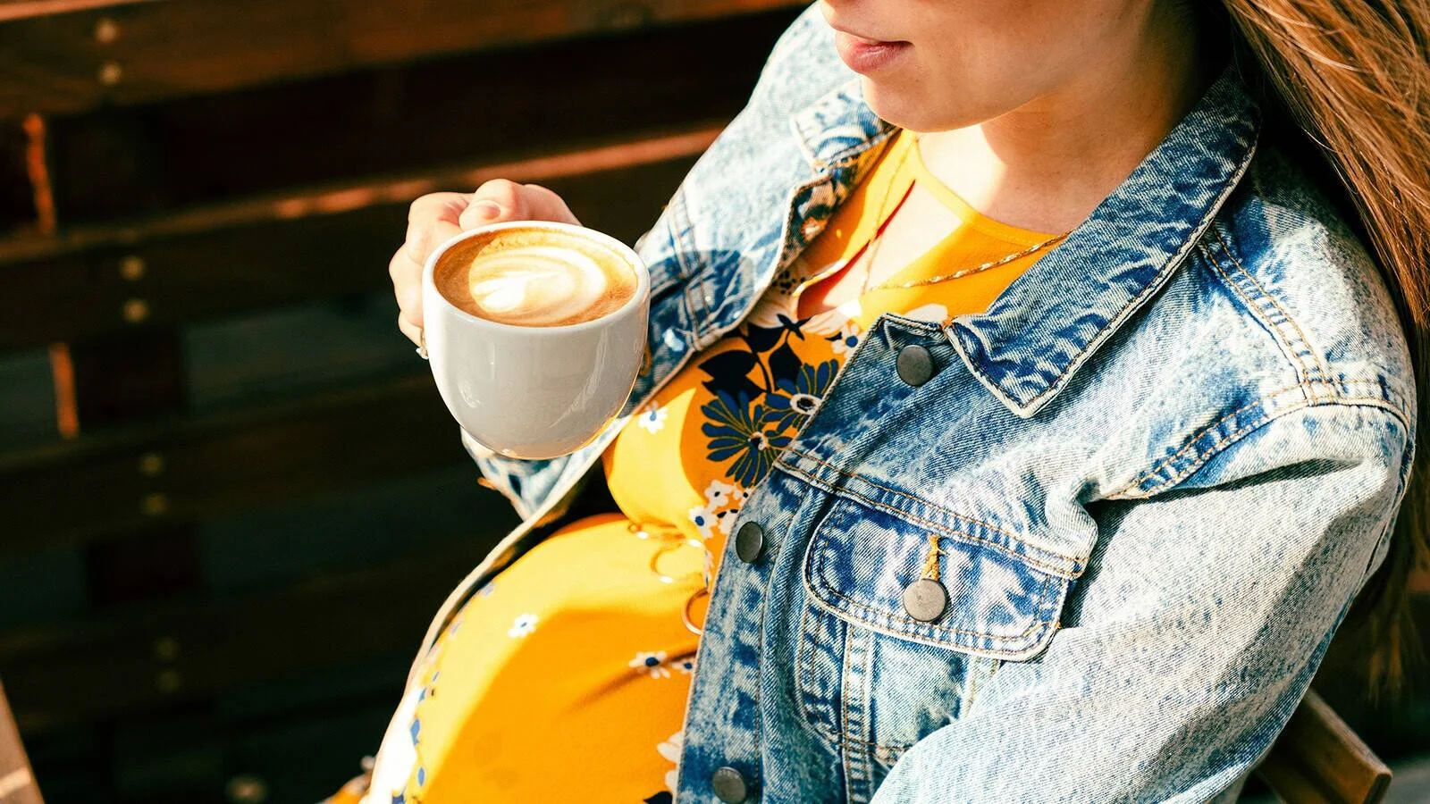 Mujer embarazada con chaqueta de jean sostiene una taza de café con leche.