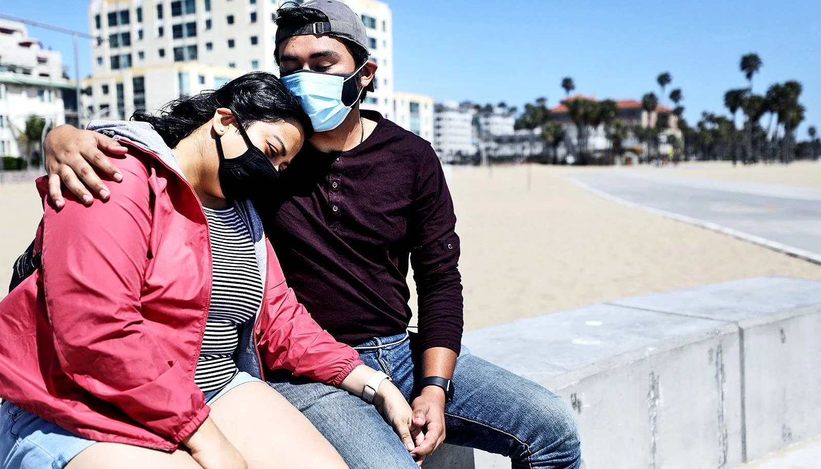 Una pareja joven con mascarillas sanitarias se abraza sentada junto a una playa, con edificios y palmeras al fondo.