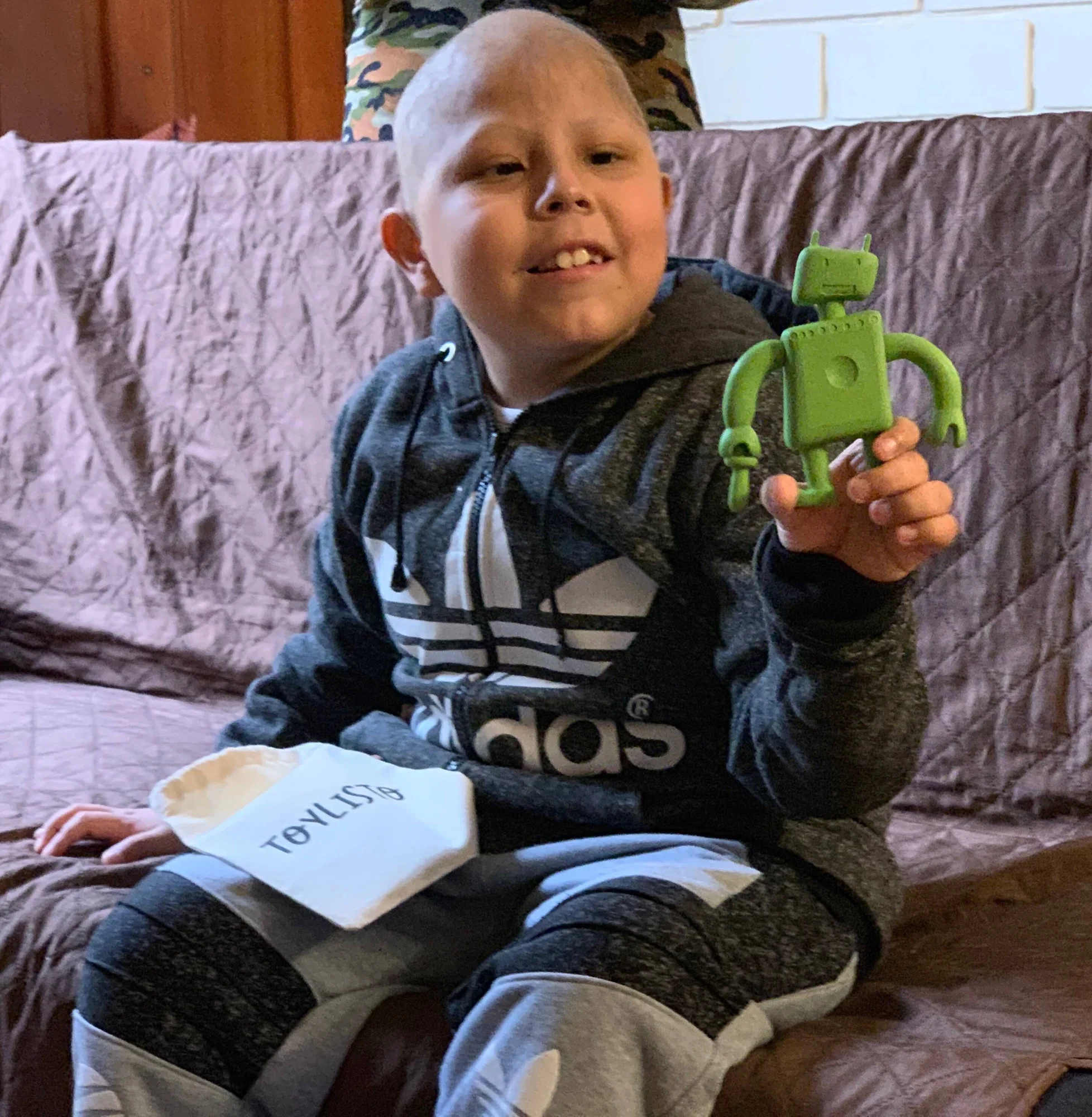 Niño con cabello muy corto sonríe sosteniendo una figura de robot verde, con una caja de juguete en el regazo.