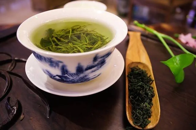Taza gaiwan de porcelana azul y blanca con té verde en infusión, junto a una cuchara de bambú con hojas de té sueltas.