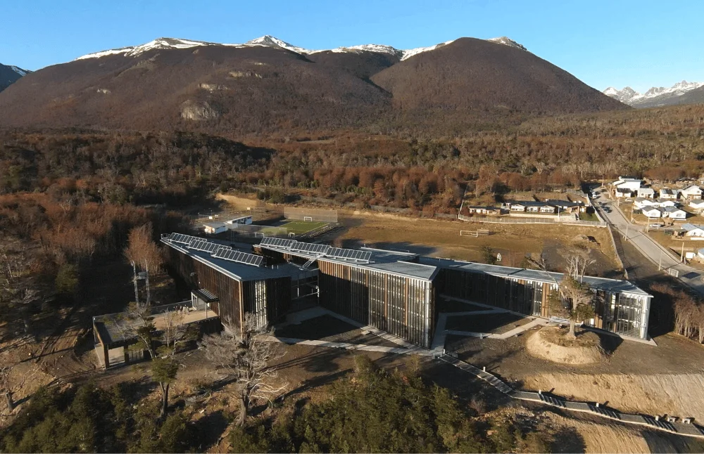 Vista aérea de un moderno edificio con paneles solares en el techo, rodeado de bosque y montañas patagónicas nevadas al fondo.