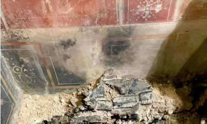 Fragmentos de madera carbonizada y ceniza en el suelo junto a una pared con frescos romanos de colores rojo y negro.