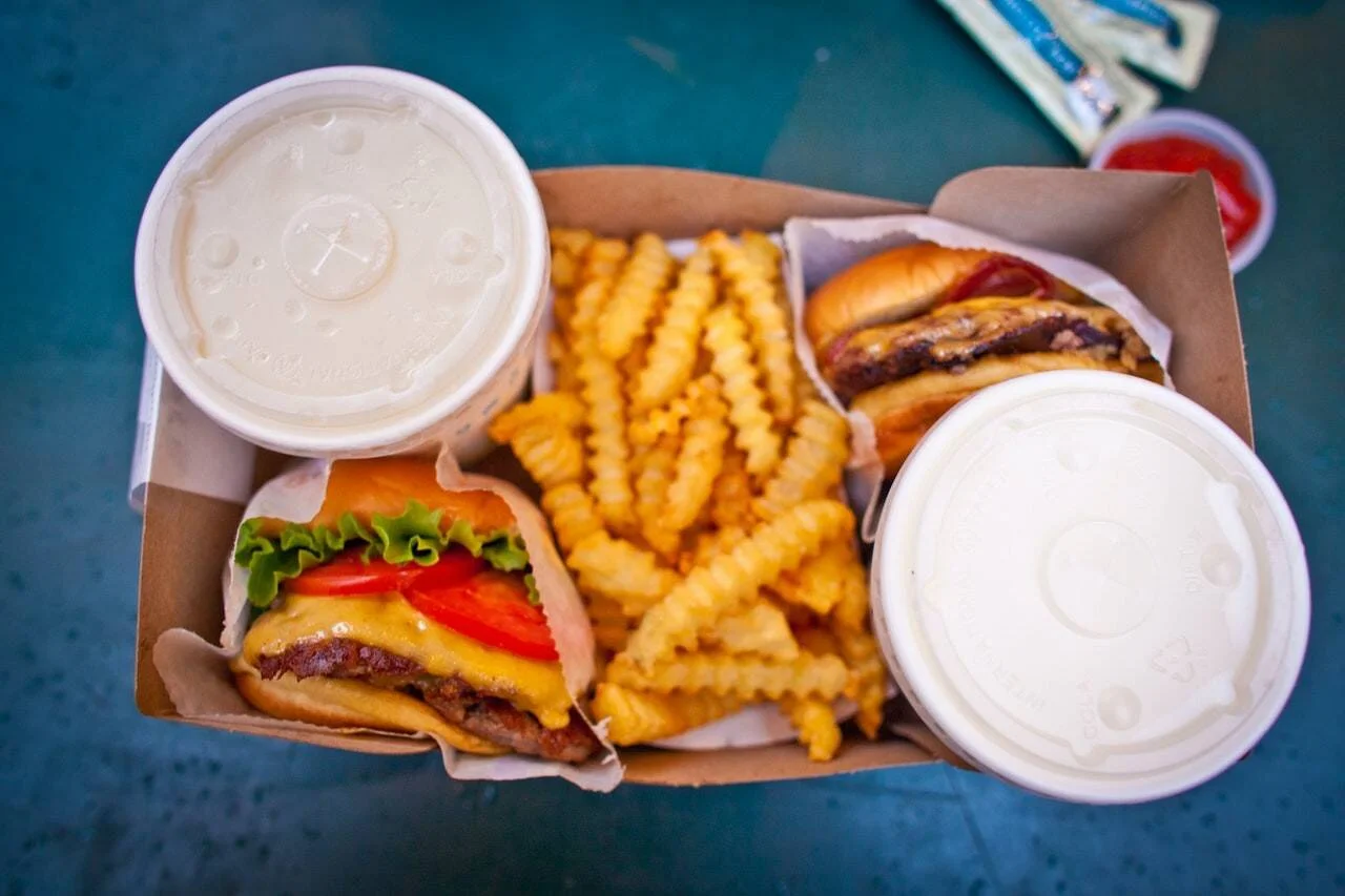 Vista cenital de una caja de comida rápida con dos hamburguesas con queso, papas fritas rizadas y dos batidos.