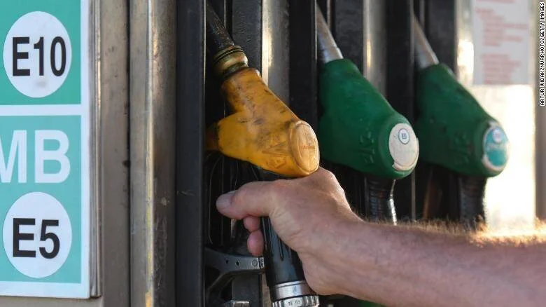 Una mano sostiene una boquilla de combustible en una estación de servicio con surtidores E5 y E10 visibles.