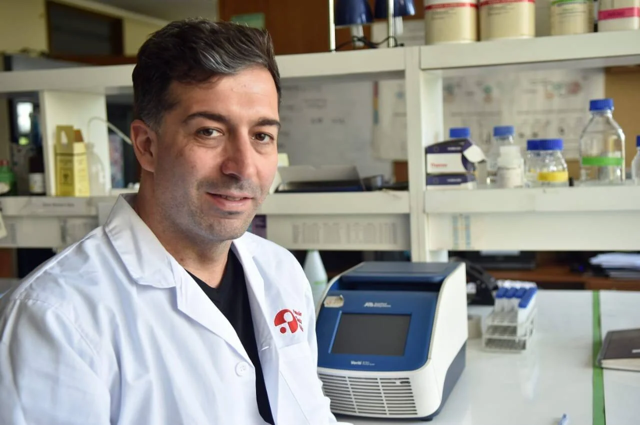 Científico con guardapolvo blanco posando en un laboratorio equipado con termociclador y reactivos en estantes.