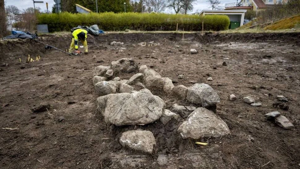 Arqueólogo trabajando en una excavación donde se aprecia una alineación de piedras en el suelo, posiblemente restos de una estructura antigua.