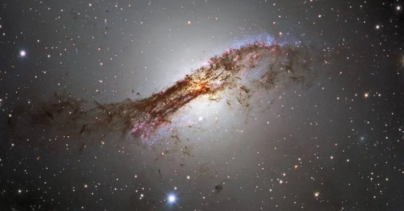 Galaxia elíptica atravesada por una banda de polvo oscuro rojizo y regiones de formación estelar azul, rodeada de estrellas.