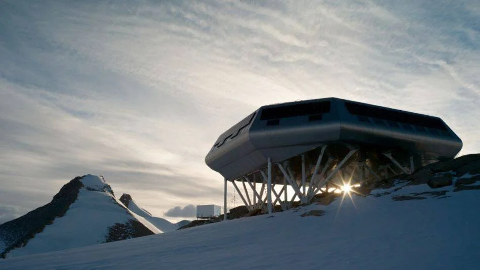Módulo habitacional de diseño aerodinámico elevado sobre pilares metálicos en un paisaje antártico nevado al atardecer.
