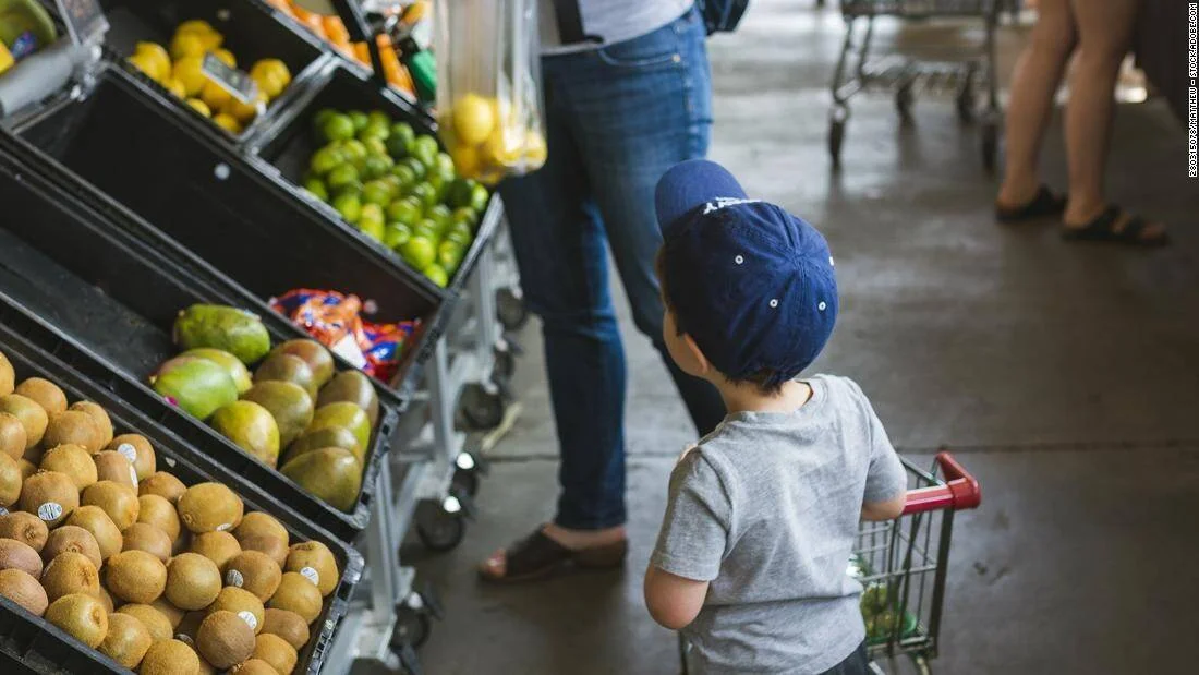 Niño con gorra azul empuja un carrito de compras frente a estantes de frutas variadas en un mercado.
