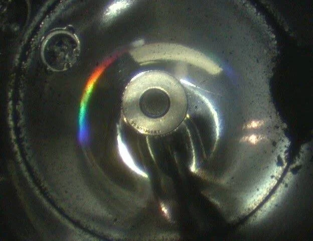 Vista interior de una pieza metálica circular con un reflejo de luz que forma un arcoíris sobre la superficie.