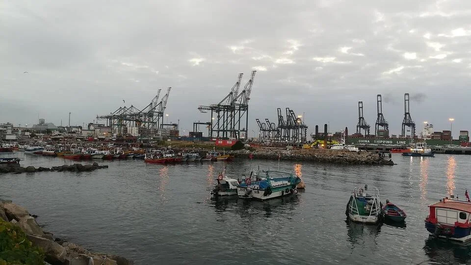 Embarcaciones pesqueras amarradas frente a un puerto de contenedores con grandes grúas bajo un cielo nublado.