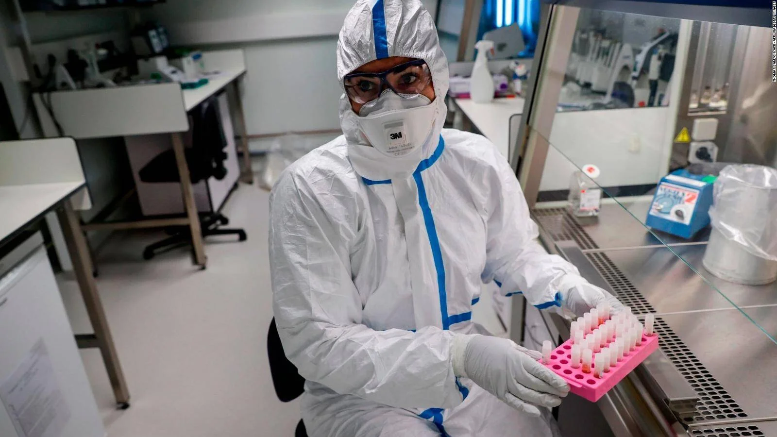 Investigador con traje de bioseguridad, mascarilla y gafas sostiene una gradilla con tubos de muestras en un laboratorio.