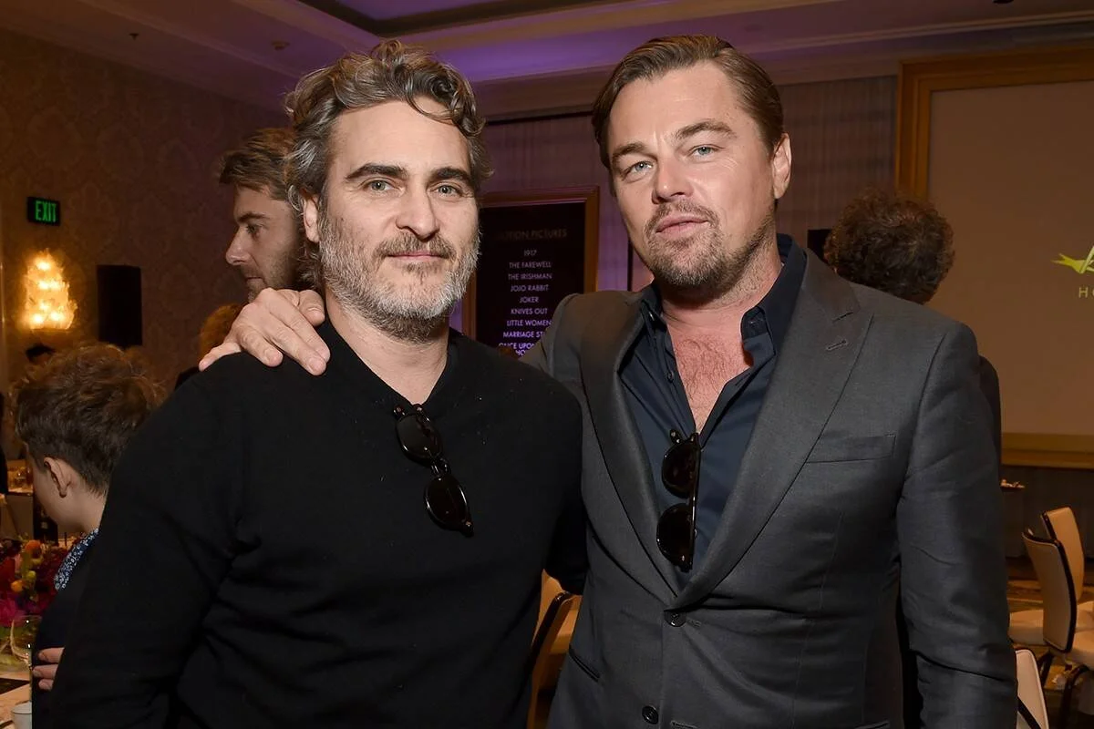 Dos hombres con trajes elegantes posan juntos en un evento de la industria cinematográfica, con una lista de películas nominadas al fondo.