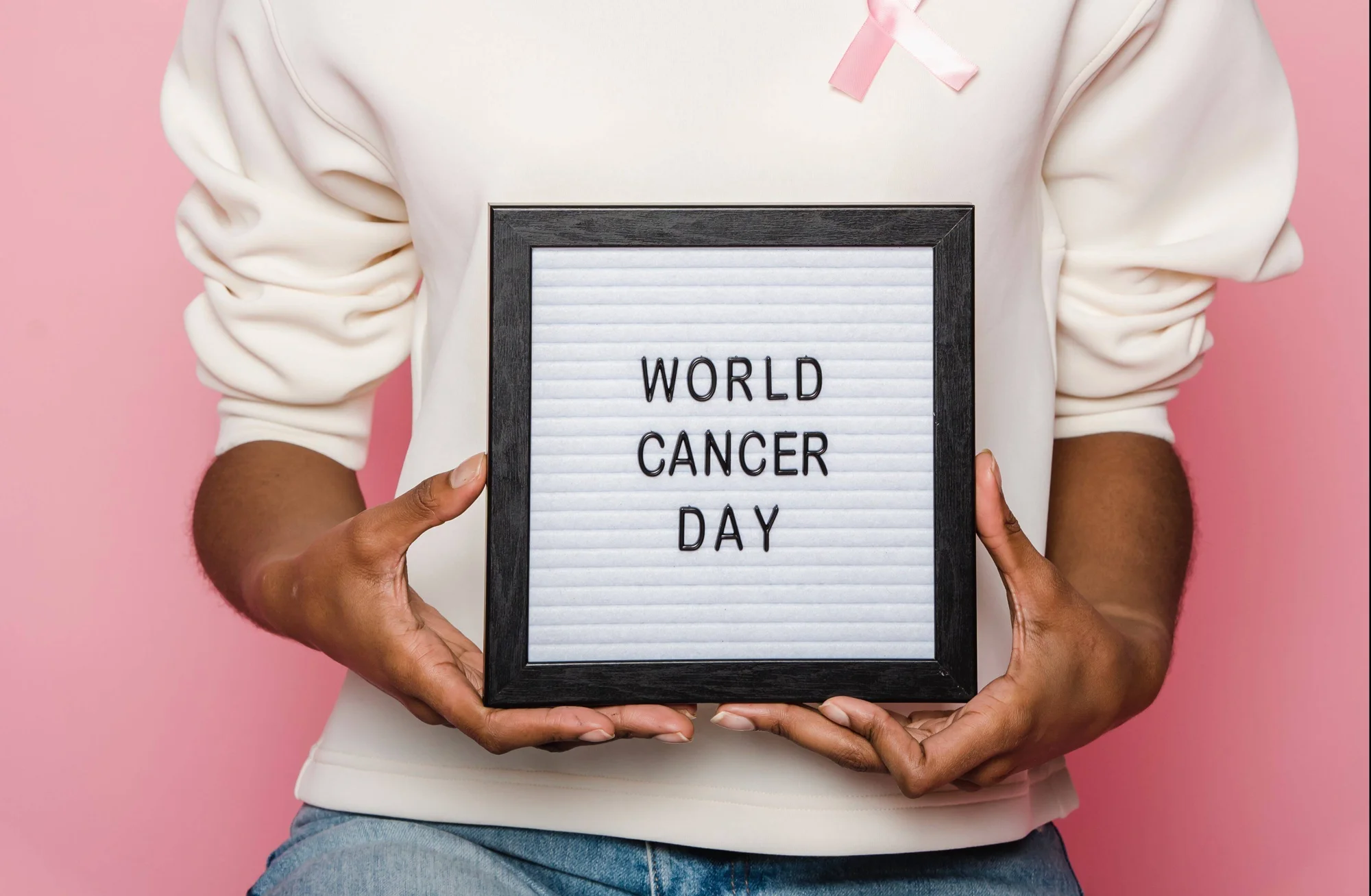 Persona con lazo rosa sostiene un cartel enmarcado con el texto 'World Cancer Day' sobre fondo rosado.