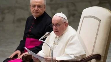 El Papa Francisco sentado en su silla, leyendo un documento durante una audiencia, acompañado por un obispo al fondo.