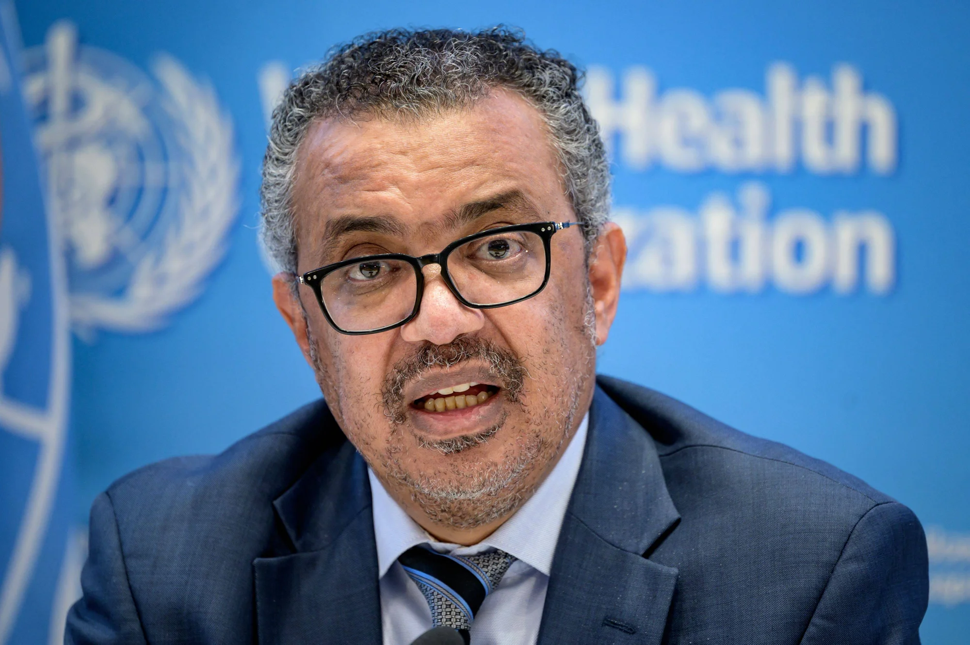 Hombre con gafas y traje azul habla en rueda de prensa ante el logo de la Organización Mundial de la Salud.