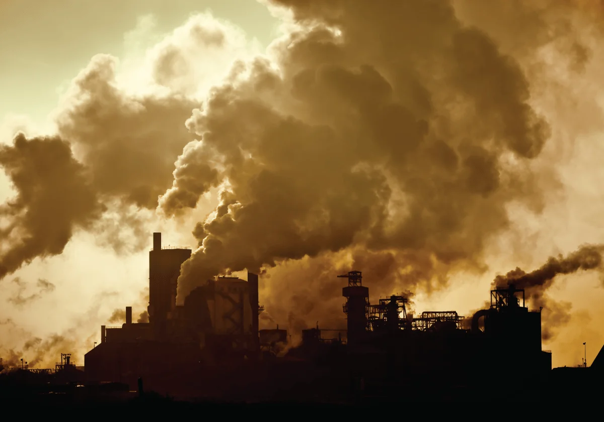 Silueta de complejo industrial con múltiples chimeneas expulsando grandes columnas de humo oscuro bajo un cielo amarillento.