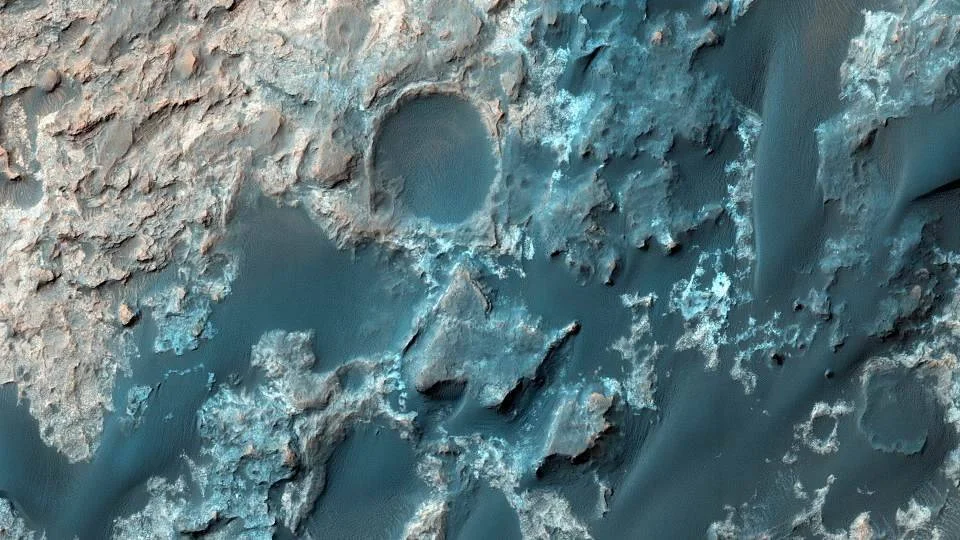 Vista aérea de la superficie de Marte con rocas claras, cráteres y terreno en tonos azules y beige captada desde órbita.