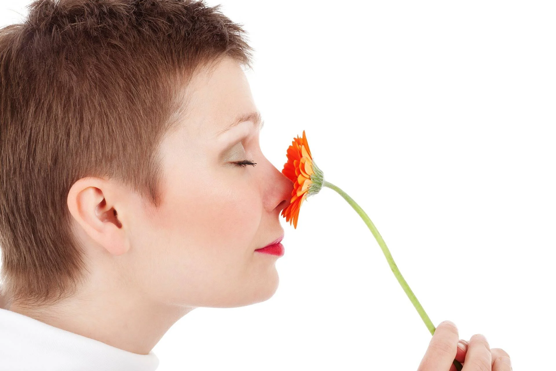 Mujer de cabello corto con los ojos cerrados oliendo una gerbera naranja sobre fondo blanco.