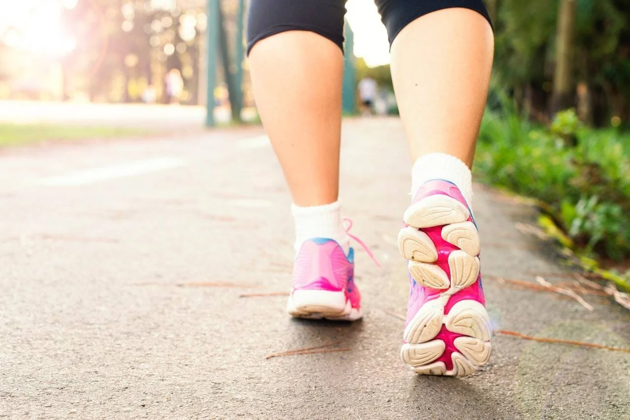 Primer plano de piernas de una persona caminando al aire libre con zapatillas deportivas rosas sobre un sendero asfaltado.
