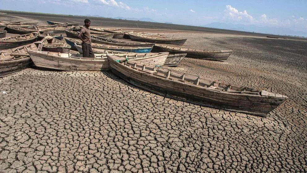 Hombre de pie entre barcas de madera abandonadas sobre el lecho agrietado y seco de un lago afectado por la sequía.