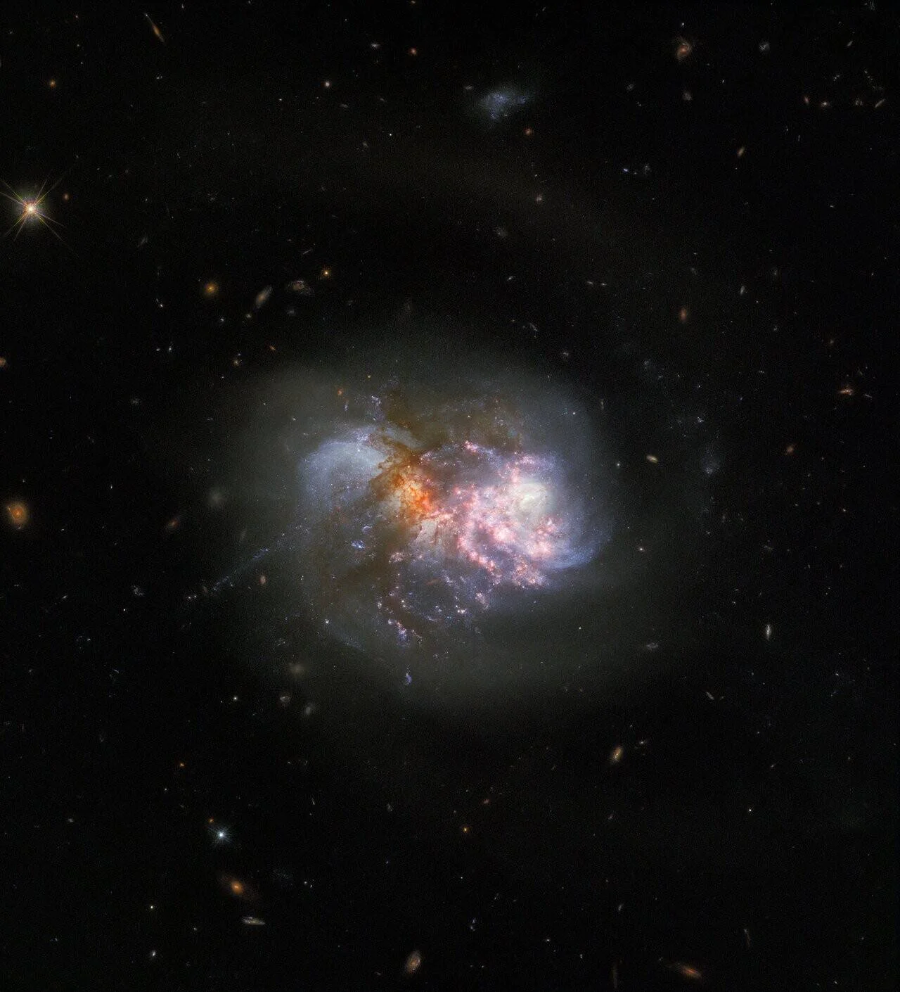 Dos galaxias en proceso de fusión, con filamentos de gas naranja y rosa y regiones de formación estelar azul, rodeadas de galaxias distantes.
