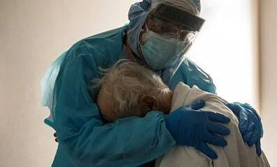 Un profesional de salud con equipo de protección completo abraza con ternura a un paciente anciano en un centro médico.