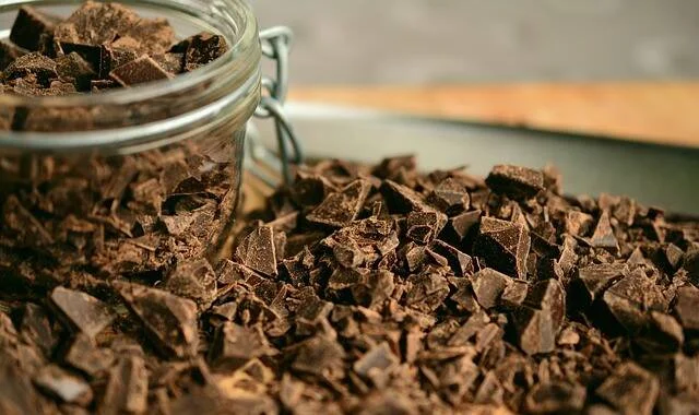 Abundantes trozos de chocolate negro picado sobre una superficie, junto a un frasco de vidrio lleno del mismo ingrediente.