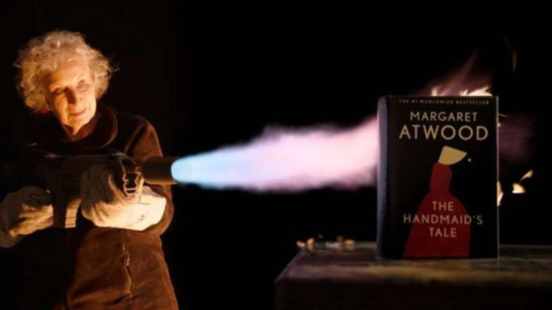 Margaret Atwood apunta un lanzallamas hacia su libro 'The Handmaid's Tale', que arde en llamas sobre una superficie.