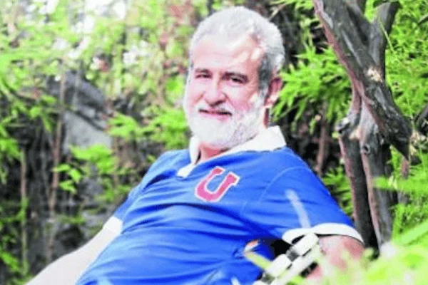 Hombre mayor de cabello y barba blancos con camiseta azul del club Universidad de Chile, sentado en entorno verde.