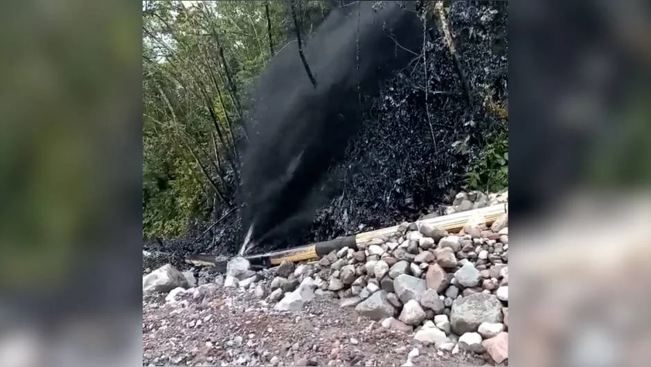 Líquido negro brota a presión desde una tubería rota junto a rocas y vegetación cubierta de residuos oscuros.