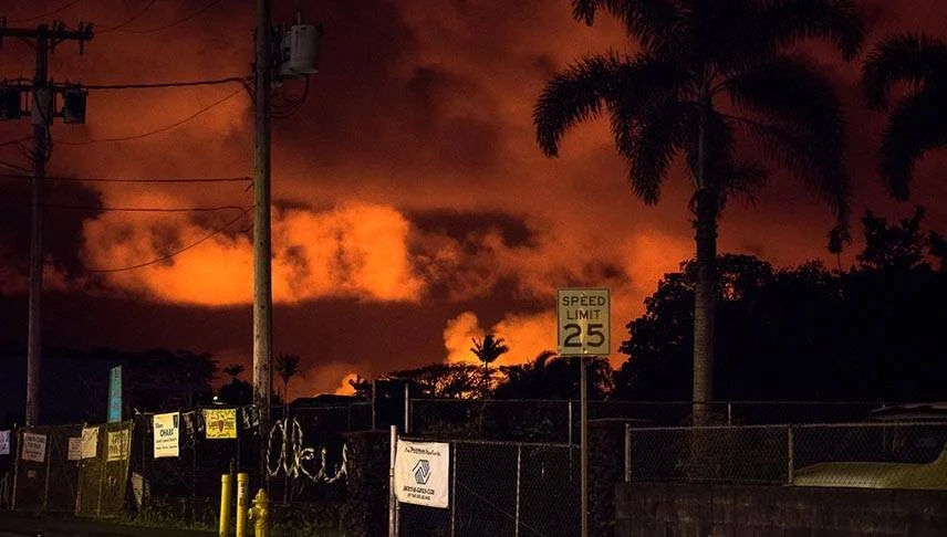 Nubes teñidas de naranja intenso por erupción volcánica iluminan el cielo nocturno sobre una calle con señal de velocidad y palmeras.