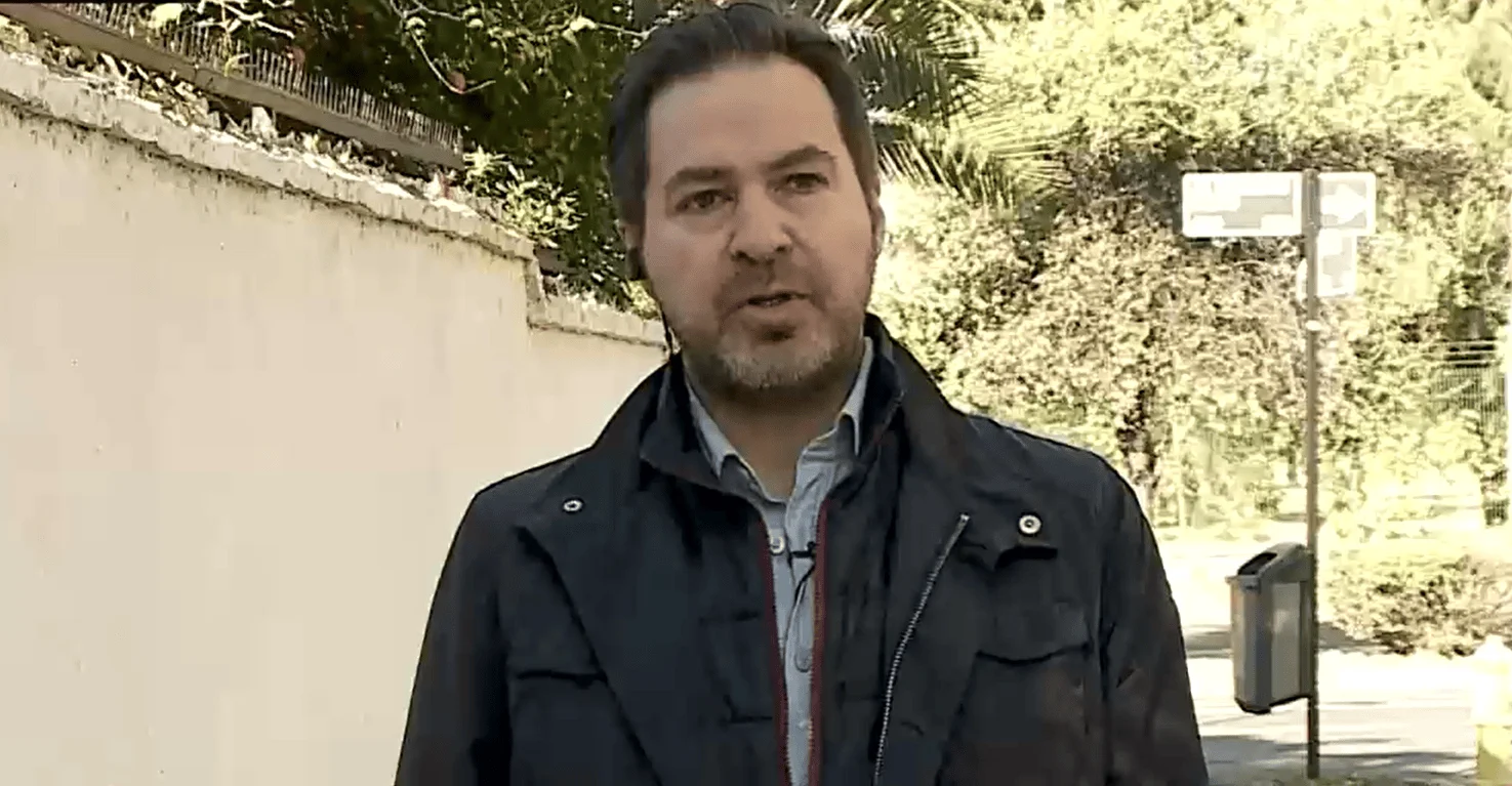 Hombre con chaqueta oscura y micrófono de solapa habla a cámara frente a un muro y vegetación en un entorno urbano.