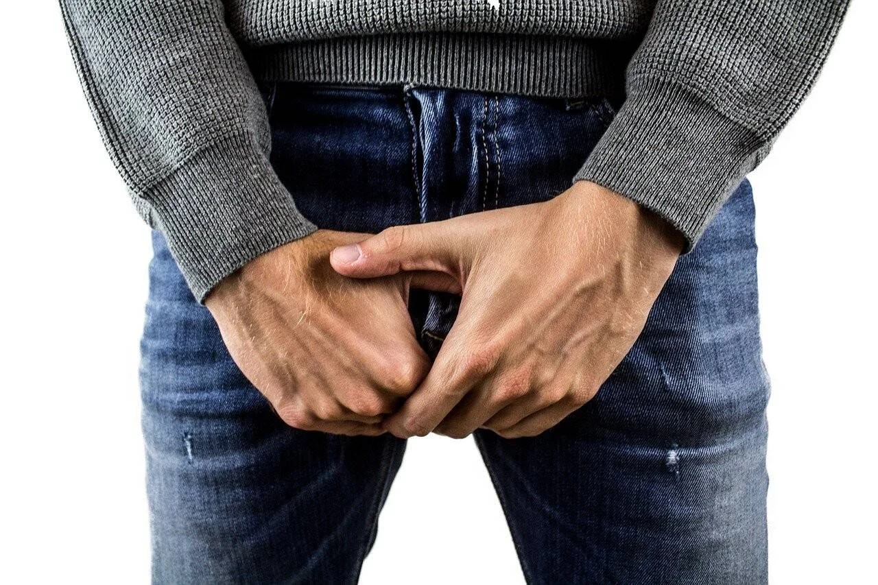 Hombre con jeans cubriendo su zona genital con ambas manos, gesto asociado a dolor o malestar en la ingle.