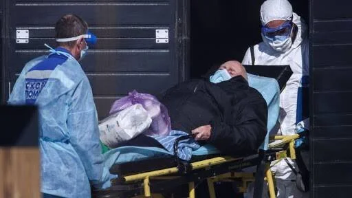 Dos trabajadores de salud con equipo de protección trasladan a un paciente en camilla hacia una instalación médica.