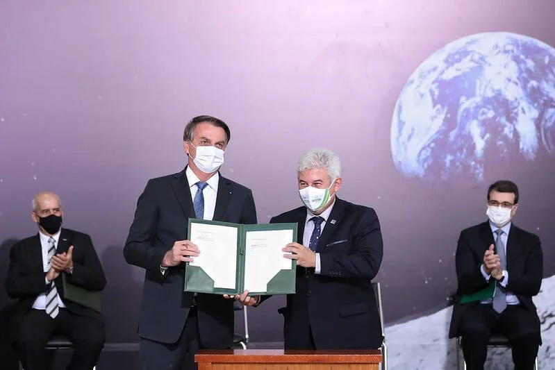 Dos funcionarios con mascarilla muestran un documento oficial firmado ante un fondo con imagen de la Tierra desde el espacio.