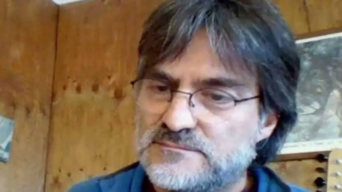Hombre de mediana edad con cabello oscuro, barba canosa y anteojos, frente a una pared de madera.