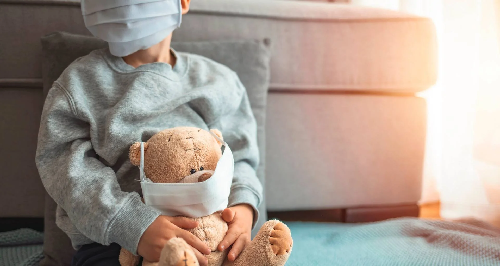 Niño con mascarilla sanitaria abraza un osito de peluche que también lleva mascarilla, sentado en el suelo de una habitación.