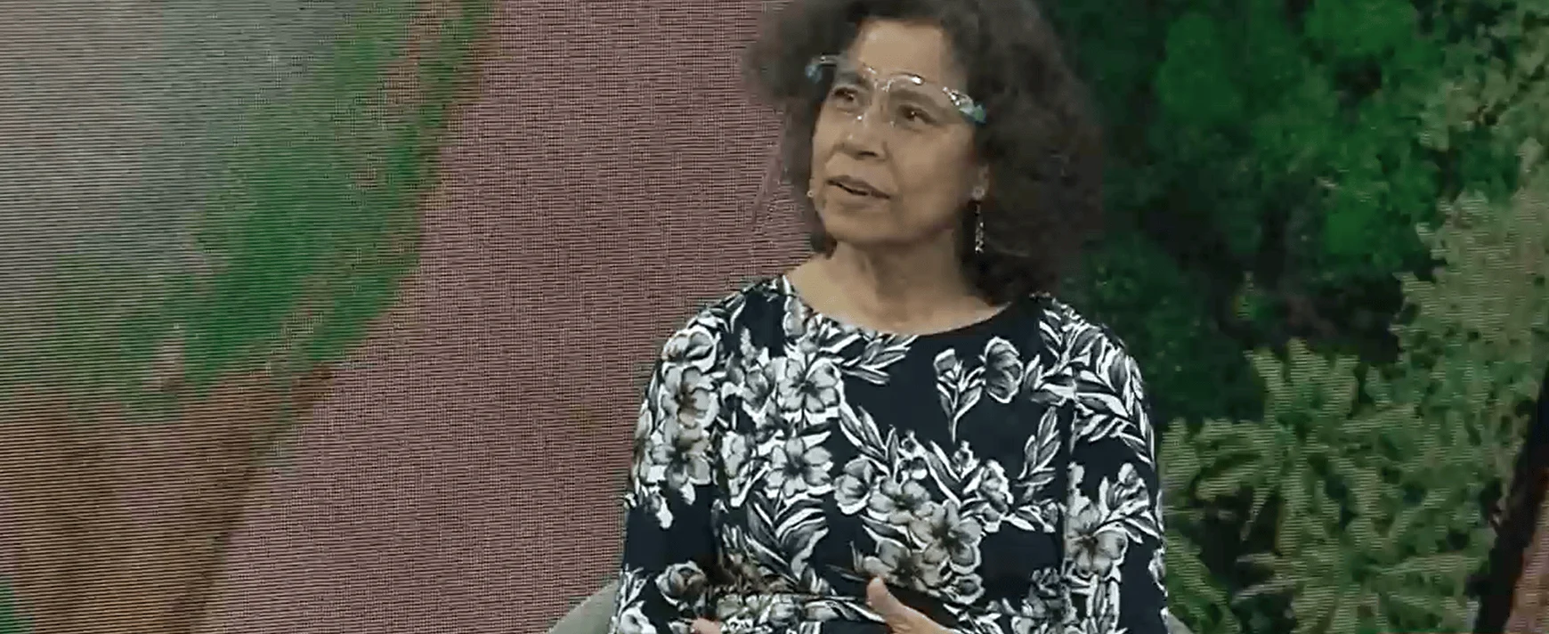 Mujer de mediana edad con lentes y blusa floral oscura habla en un escenario con fondo vegetal.