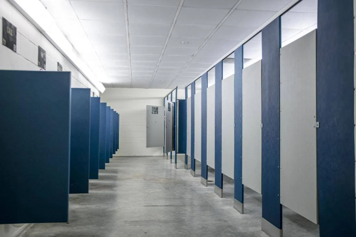 Interior de un baño público amplio con múltiples cubículos de color azul y blanco, piso de cemento y techo iluminado.