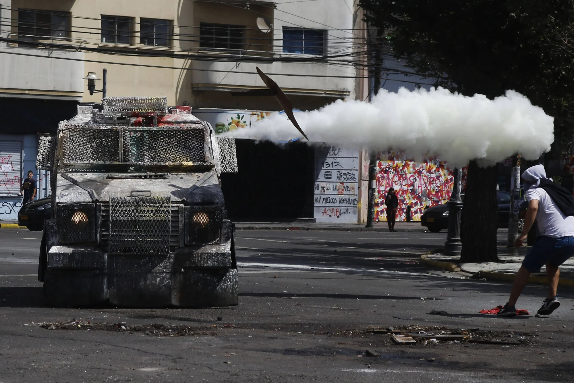 Vehículo blindado antidisturbios dispara gas lacrimógeno mientras un manifestante encapuchado huye en una calle urbana.