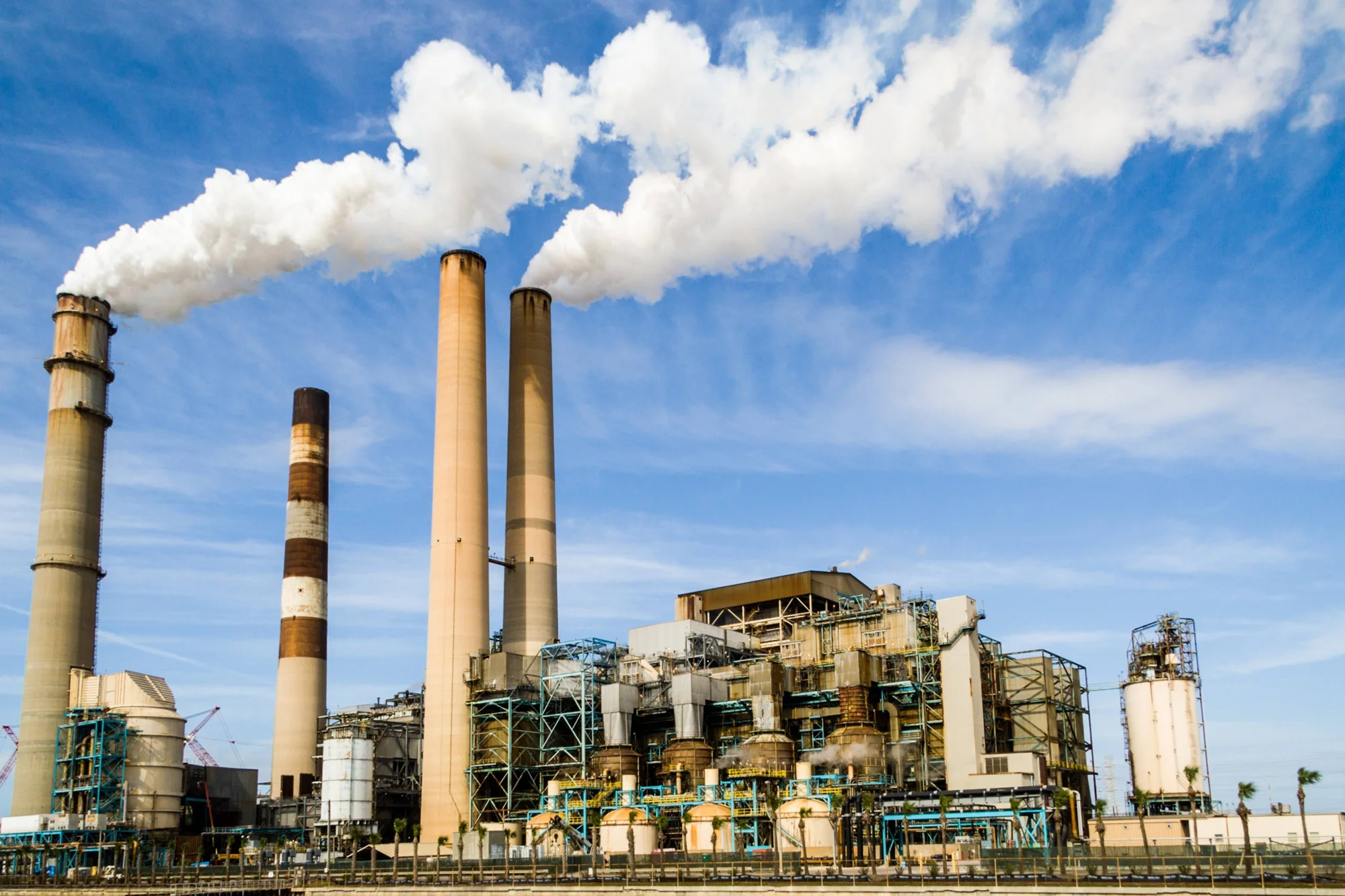 Central termoeléctrica con cuatro chimeneas expulsando grandes columnas de humo blanco hacia un cielo azul.