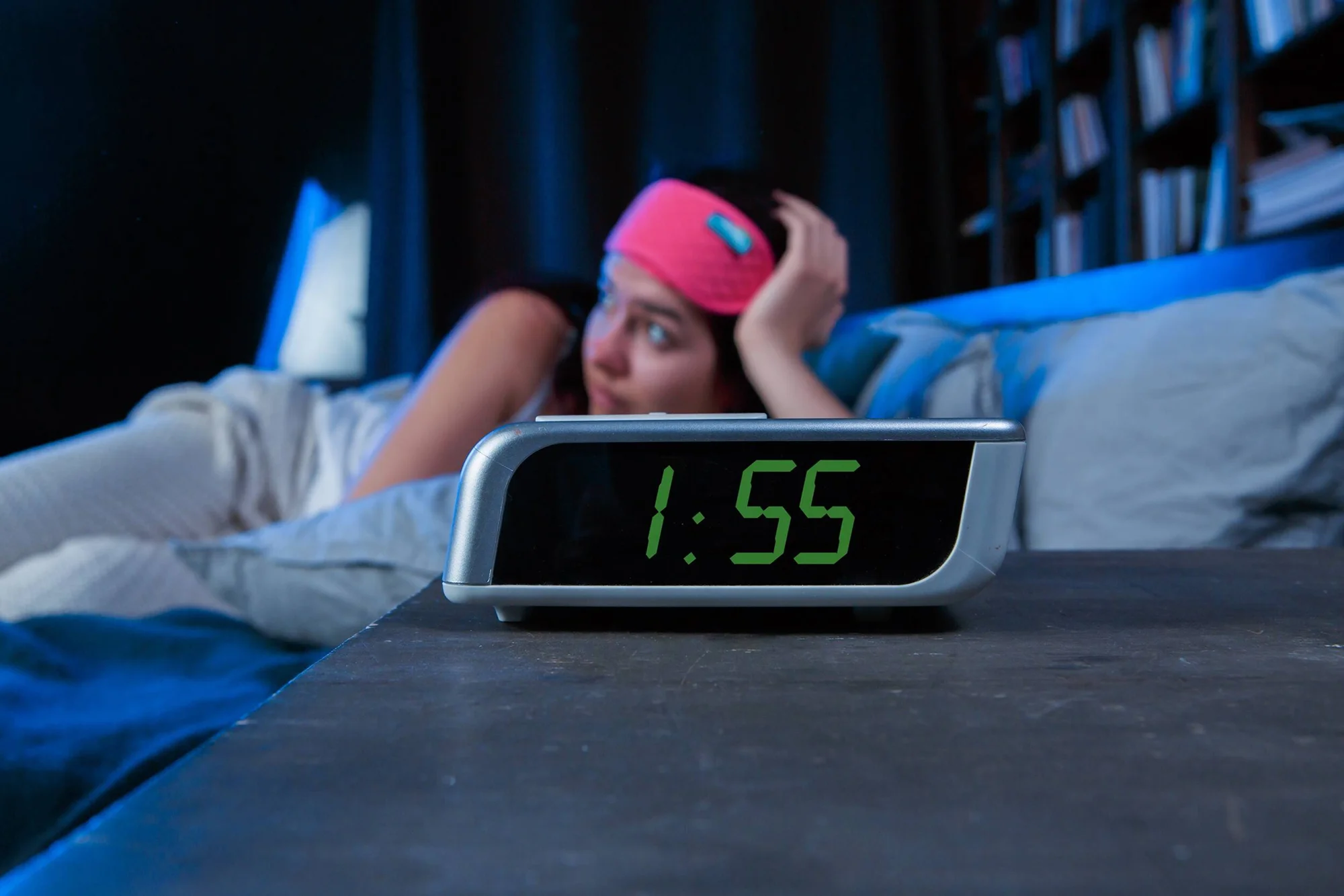 Mujer acostada con antifaz rosa mira el reloj despertador que marca la 1:55, reflejando insomnio nocturno.