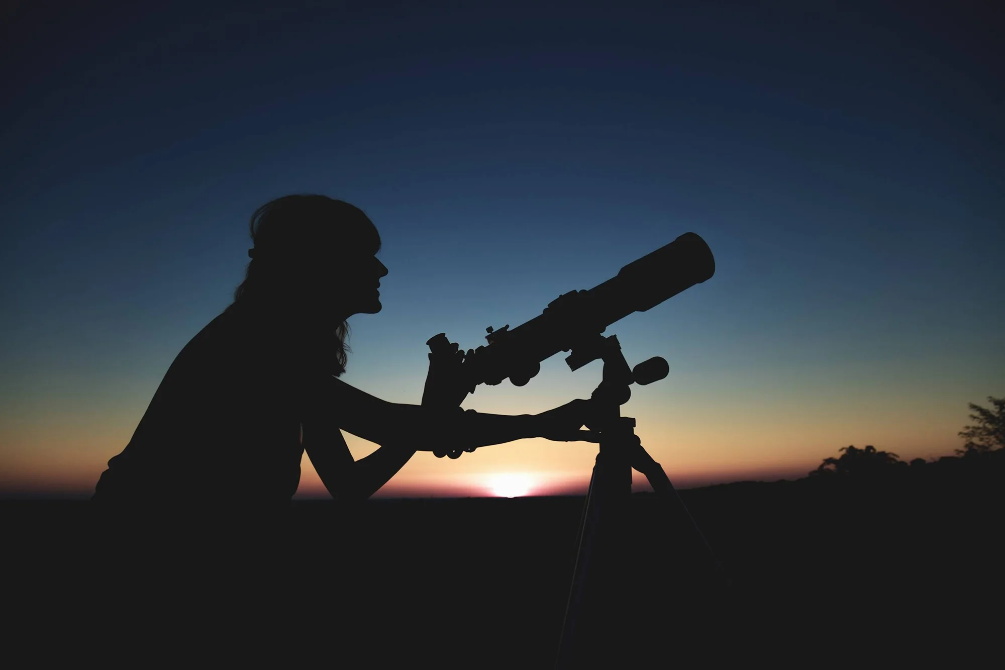 Silueta de una persona observando el cielo con un telescopio sobre trípode durante el crepúsculo.