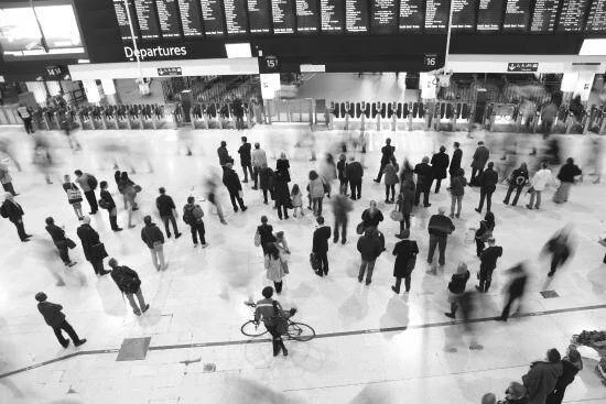 Vista aérea en blanco y negro de pasajeros aguardando en el vestíbulo de una estación de tren bajo los paneles de salidas.