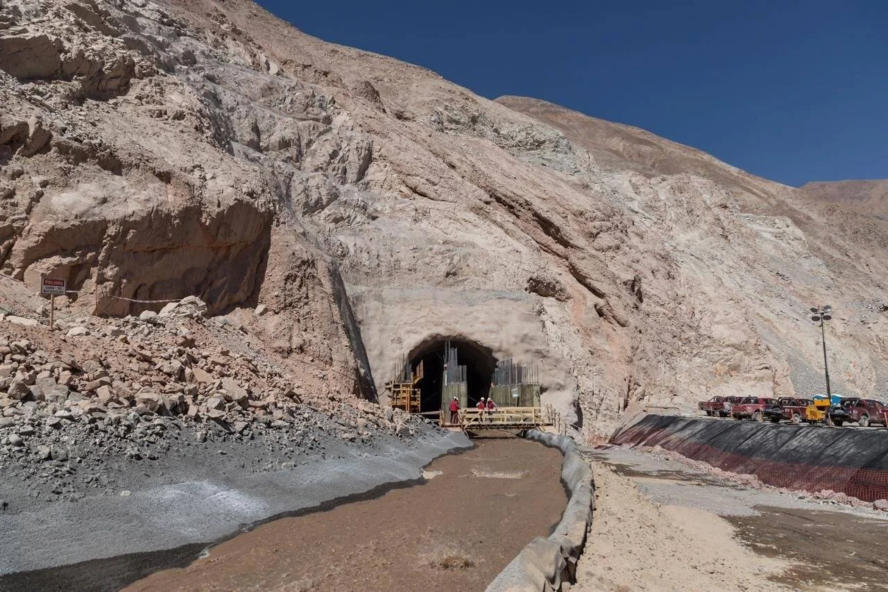 Boca de un túnel en construcción excavado en una montaña rocosa árida, con trabajadores y maquinaria en el acceso.