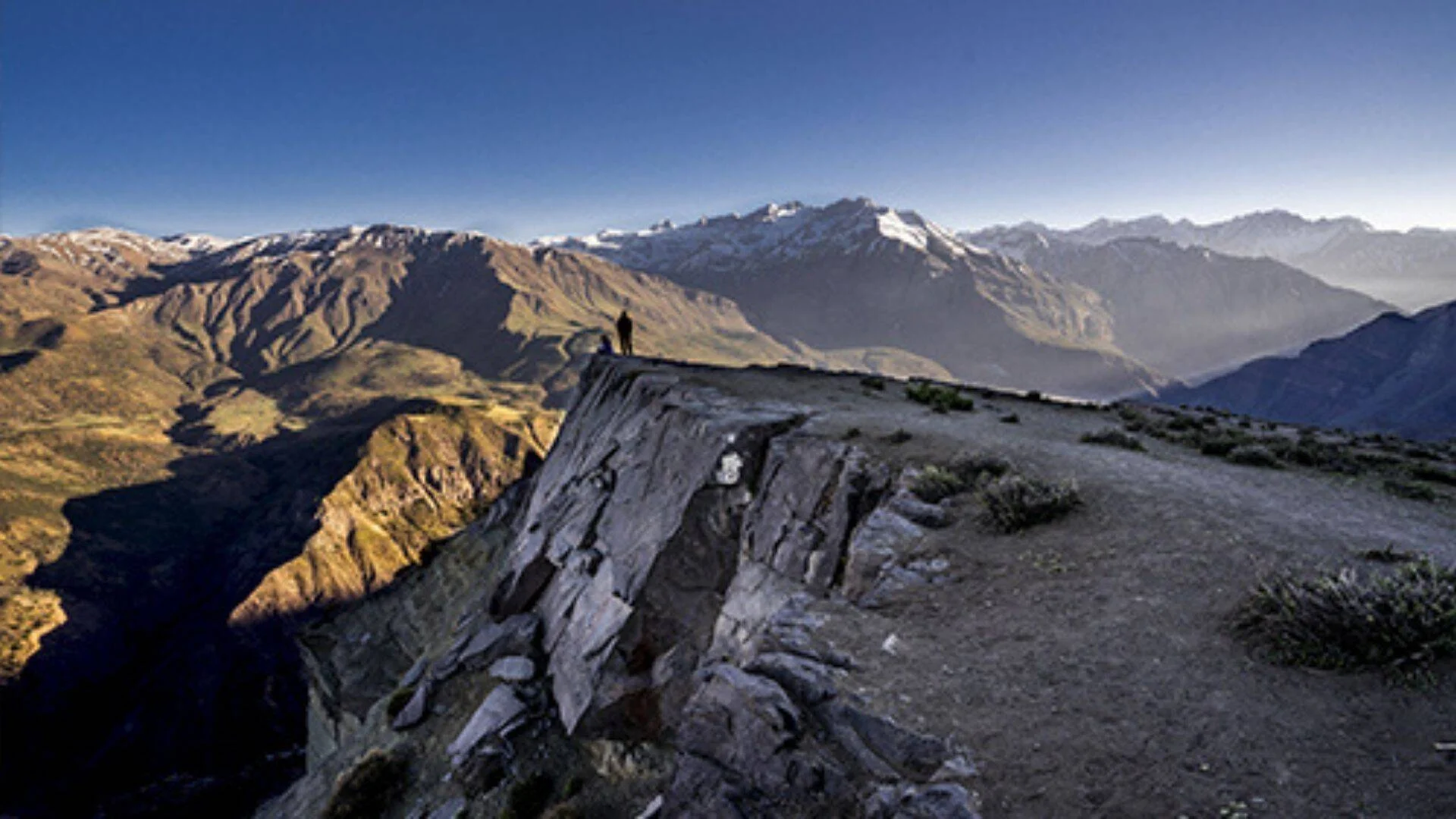 Una persona de pie al borde de un acantilado rocoso contempla una extensa cordillera con cumbres nevadas bajo un cielo azul despejado.