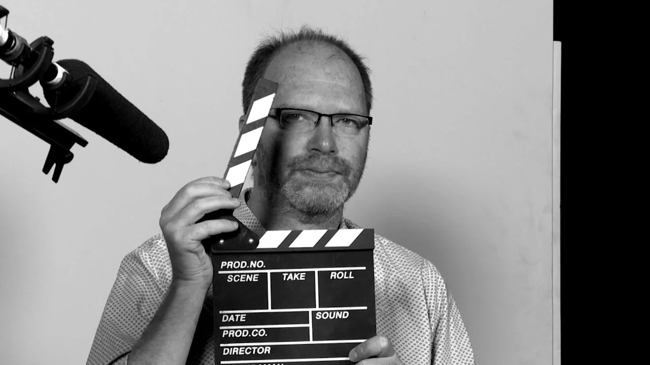 Hombre con gafas sostiene una claqueta de filmación frente a su rostro junto a un micrófono de estudio, en blanco y negro.