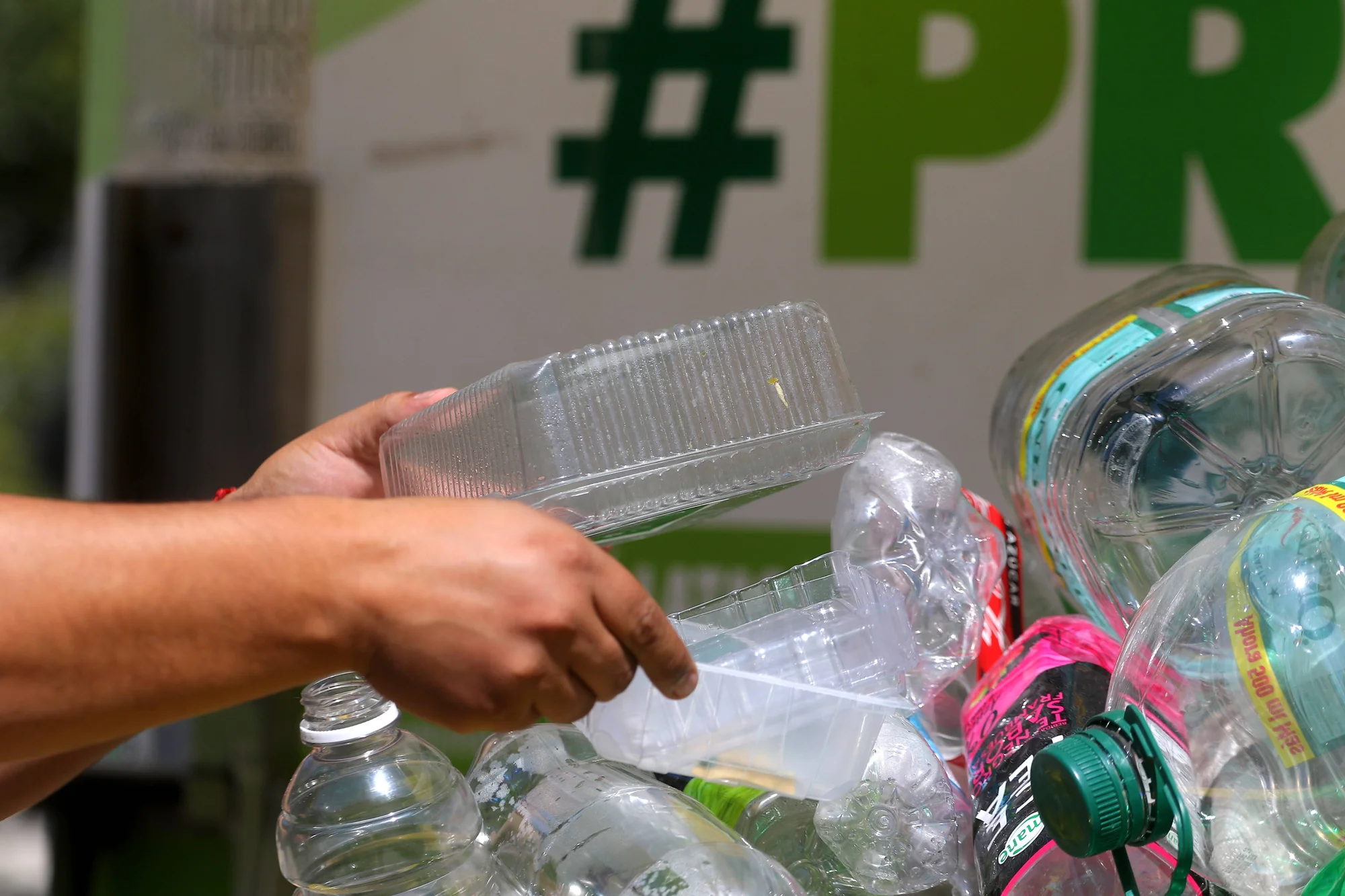 Una mano deposita un envase plástico transparente sobre un conjunto de botellas PET en un punto de reciclaje.