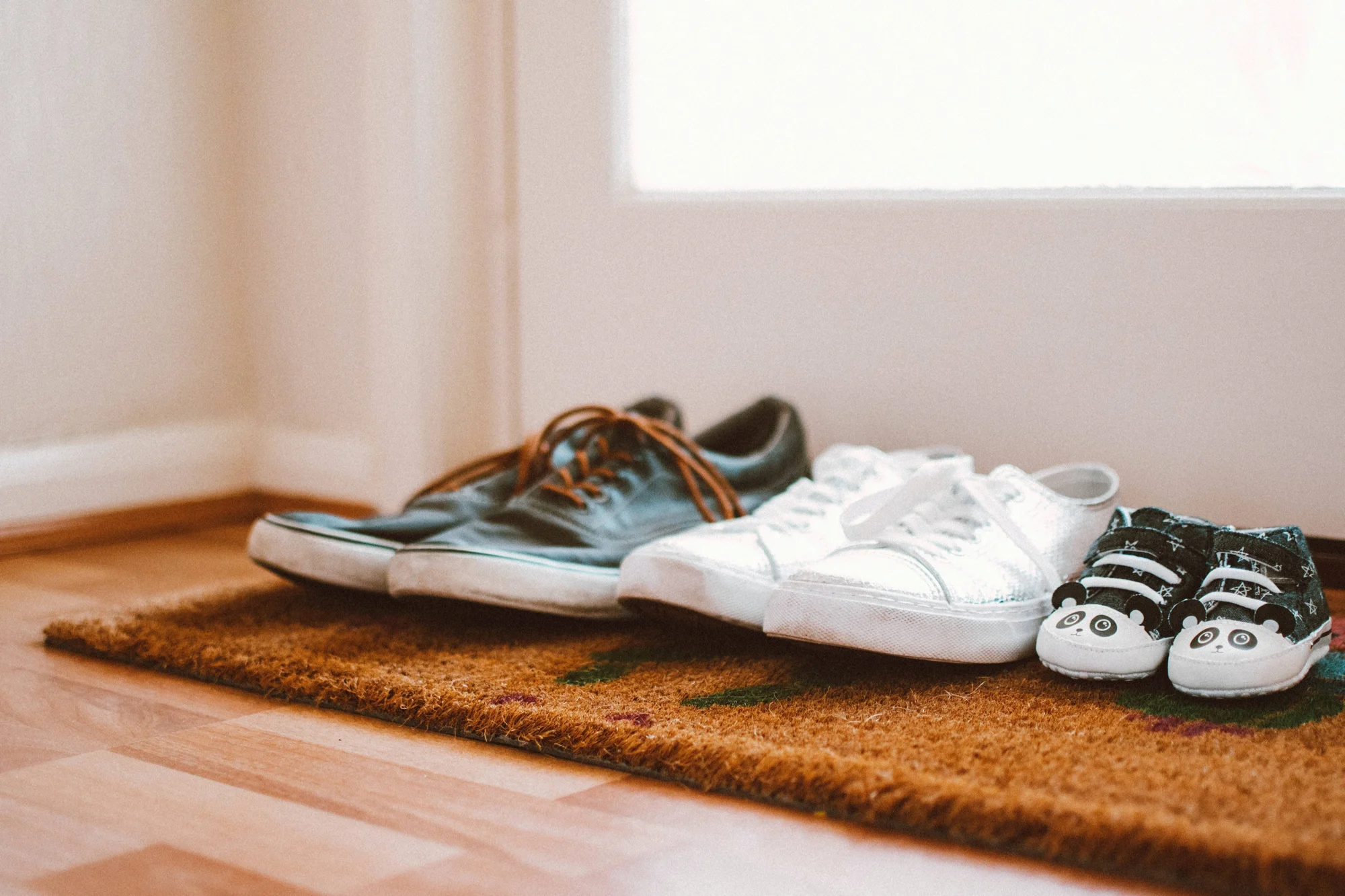Tres pares de zapatillas de distintos tamaños —adulto, joven y bebé con diseño de panda— alineados sobre un felpudo de yute en la entrada de un hogar.